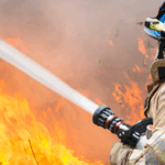 A firefighter wearing a blue helmet uses a fire hose to fight a large flame.