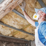 Worker wearing a hard hat, gloves, and filtration mask inspects yellow insulation between wooden ceiling beams.
