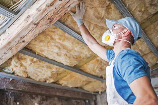 Worker wearing a hard hat, gloves, and filtration mask inspects yellow insulation between wooden ceiling beams.