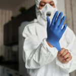 Worker in white protective coveralls, respirator mask, and blue gloves preparing for cleanup in an indoor room.